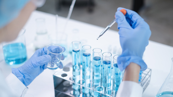 Laborant u bijelom kutu s plavim rukavicama drži čašicu i pipetu. Ispred njega je red cijevi za ispitivanje s plavom tekućinom, a na radnoj površini su različite labotatorijske boce s plavim tekućinama.