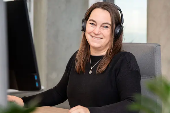 Žena sjedi za radnim stolom s računalom, nosi slušalice i osmjehuje se. Ima dužu smešnu kosu i obučena je u crnu majicu. U pozadini su elementi modernog ureda.