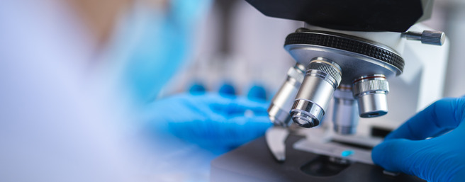U fokusu je mikroskop s plavim rukavicama koje ga koriste. Vidljive su leće mikroskopa i rukopisna priprema ispod. Laboratorijski ambijent uključuje blurirane pozadine.
