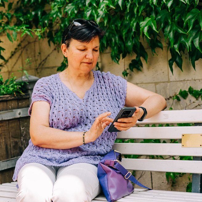 Žena sjedi na klupi u parku, nosi svijetlu ljubičastu majicu i svijetle hlače. U ruci drži pametni telefon i gleda u njega. Iza nje se vidi gusta zelena vegetacija.