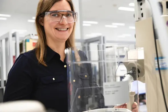 Žena u laboratorijskom okruženju nosi zaštitne naočale i osmijeh dok stoji uz laboratorijsku opremu. Iza nje su police s istraživačkim materijalima.