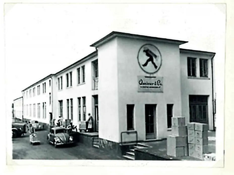 Zgrada tvrtke s dugačkom fasadom, prekrivenom prozorima i logotipom na prednjoj strani. Pored zgrade su parkirani automobili, a ispred se nalaze kutije. Fotografija je u crno-bijeloj boji.