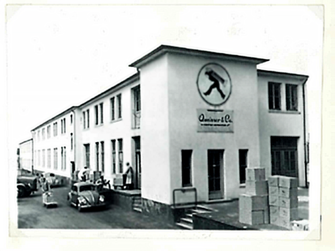 Zgrada tvrtke s dugačkom fasadom, prekrivenom prozorima i logotipom na prednjoj strani. Pored zgrade su parkirani automobili, a ispred se nalaze kutije. Fotografija je u crno-bijeloj boji.