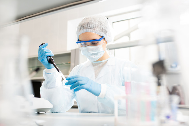 Laborant u bijelom kutu i zaštitnoj opremi, s plavim rukavicama, koristi pipetu za izvođenje eksperimenata u laboratoriju. U pozadini se vide laboratorijski uređaji i posude.