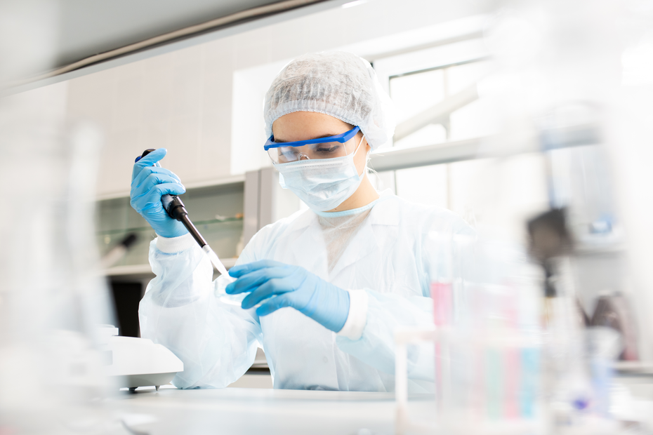 Laborant u bijelom kutu i zaštitnoj opremi, s plavim rukavicama, koristi pipetu za izvođenje eksperimenata u laboratoriju. U pozadini se vide laboratorijski uređaji i posude.