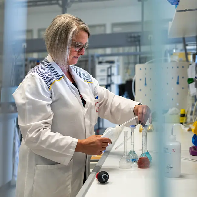 Žena u laboratorijskoj halji miješa tekućine u staklenim posudama. Okružena je laboratorijskom opremom i alatima, izgledajući fokusirano dok izvodi eksperimente.
