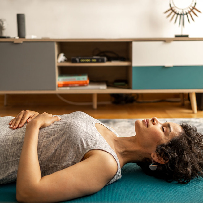 Žena leži na podu na yoga prostirci, opuštena, s rukama uz tijelo. Ima kratku tamnu kosu i nosi sivu majicu bez rukava. U pozadini se vide drveni pod i namještaj s policama.