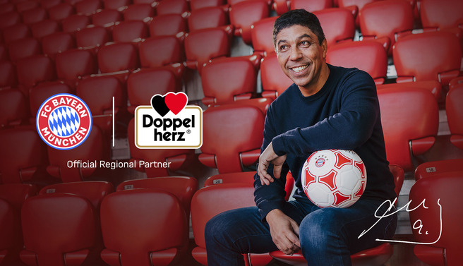 Osoba sjedi na stadionu s crvenim sjedalicama i drži nogometnu loptu. U pozadini su logotipi FC Bayern Münchena i Doppelhauta kao službenog regionalnog partnera. Osoba se smiješi i izgleda veselo.