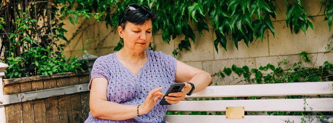 Žena s kratkom kosom sjedi na klupi pod krošnjom, drži mobilni telefon i gleda u njega. Ima na sebi ljubičastu majicu s uzorkom i sunčane naočale. U pozadini su biljke koje se penju uz zid.